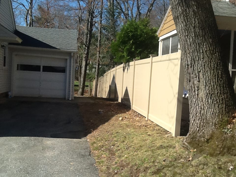 A wooden fence stands beside a gravel driveway, with a leafy tree providing shade in the background.
