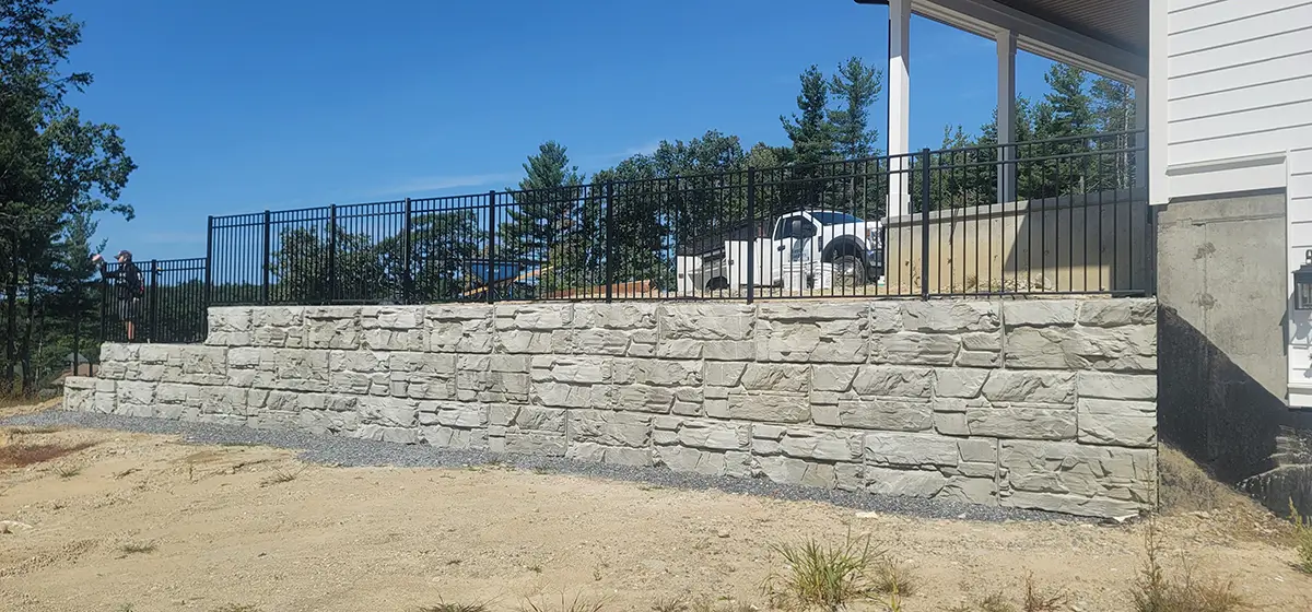 A construction worker builds a retaining wall in front of a house, with tools and materials visible on the ground.
